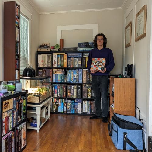 Isaac holding the board game, Azul, while standing in front of multiple bookshelves that house his 100+ boardgames.